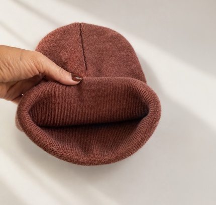 A hand holds a ribbed knit beanie in a dusty rose-brown color, showing the folded cuff and center seam against a clean white background with soft diagonal shadows.