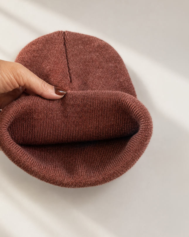 A hand holds a ribbed knit beanie in a dusty rose-brown color, showing the folded cuff and center seam against a clean white background with soft diagonal shadows.