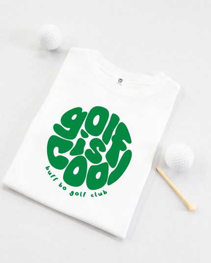 Folded white kids' t-shirt with bold green text that reads "golf is cool" in a playful, rounded font and "buff bo golf club" underneath, displayed on a light background with two white golf balls and a wooden tee.

