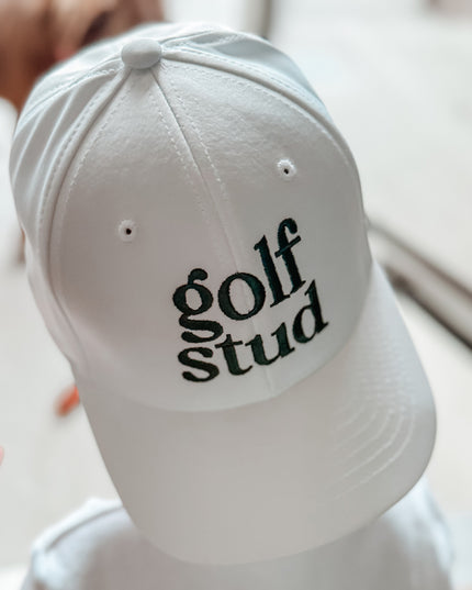 Child wearing a white baseball cap with bold green embroidered 'golf stud' text, casual and playful golf hat perfect for kids and youth golfers, photographed indoors with soft natural light.