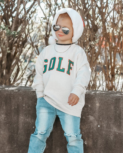 Child wearing a 'GOLF' sweatshirt, sunglasses, and ripped jeans against a natural background.