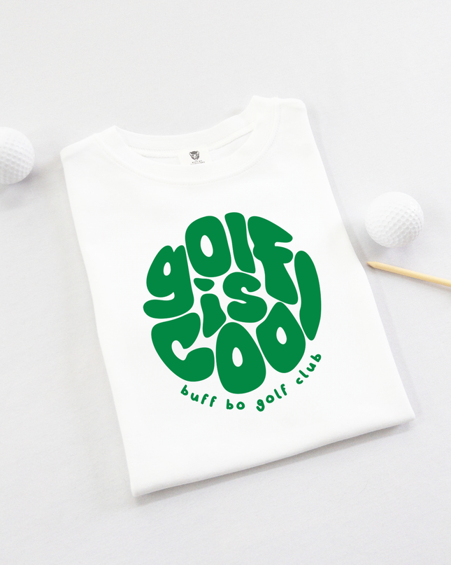 Folded white kids' t-shirt with bold green text that reads "golf is cool" in a playful, rounded font and "buff bo golf club" underneath, displayed on a light background with two white golf balls and a wooden tee.
