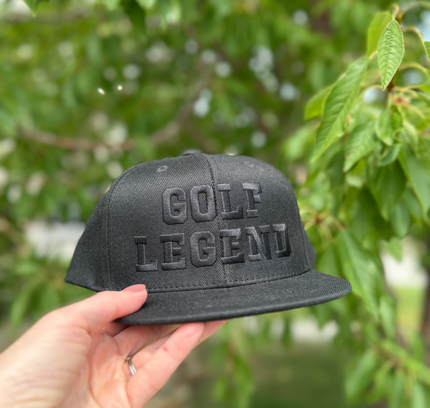 Black cap with 'GOLF LEGEND' text held by a hand against a blurred green background