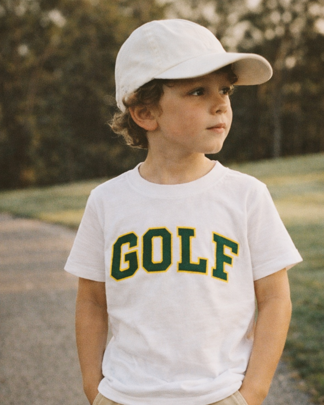 Young boy wearing a white t-shirt with 'GOLF' printed on it, standing outdoors.