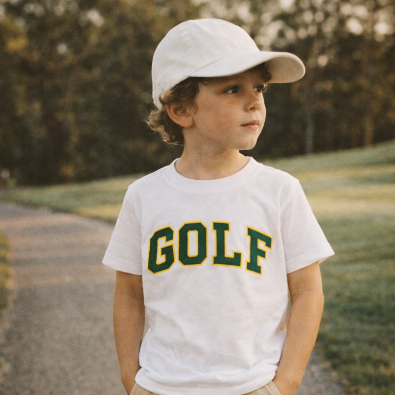 Young boy wearing a white t-shirt with 'GOLF' printed on it, standing outdoors.