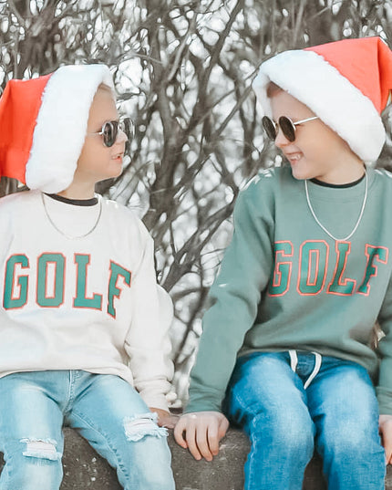 Two children wearing Santa hats and 'GOLF' sweatshirts sitting on a ledge with bare trees in the background.