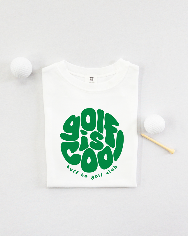 Folded white kids' t-shirt with bold green text that reads "golf is cool" in a playful, rounded font and "buff bo golf club" underneath, displayed on a light background with two white golf balls and a wooden tee.