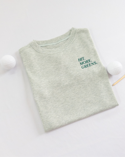 Light green kids golf t-shirt with minimalist text reading 'Hit. More. Greens.' in dark green, displayed flat lay with golf balls and a wooden tee, perfect for young golf enthusiasts.