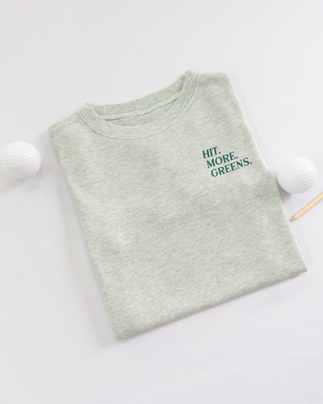 Light green kids golf t-shirt with minimalist text reading 'Hit. More. Greens.' in dark green, displayed flat lay with golf balls and a wooden tee, perfect for young golf enthusiasts.