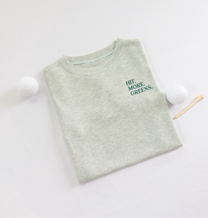 Light green kids golf t-shirt with minimalist text reading 'Hit. More. Greens.' in dark green, displayed flat lay with golf balls and a wooden tee, perfect for young golf enthusiasts.