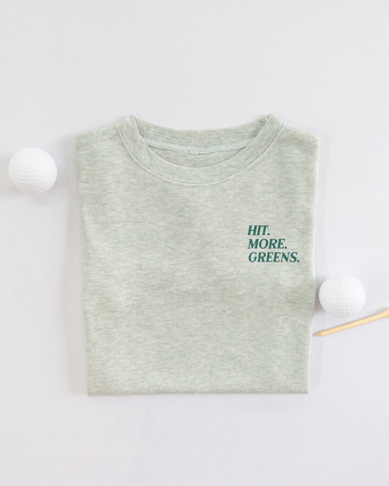 Light green kids golf t-shirt with minimalist text reading 'Hit. More. Greens.' in dark green, displayed flat lay with golf balls and a wooden tee, perfect for young golf enthusiasts.