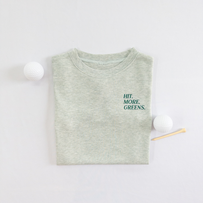 Light green kids golf t-shirt with minimalist text reading 'Hit. More. Greens.' in dark green, displayed flat lay with golf balls and a wooden tee, perfect for young golf enthusiasts.