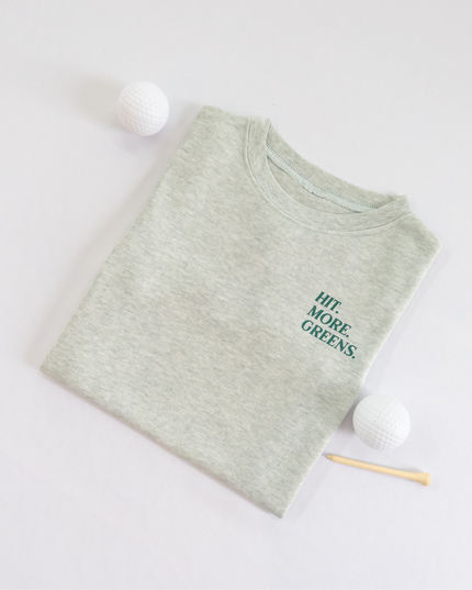 Light green kids golf t-shirt with minimalist text reading 'Hit. More. Greens.' in dark green, displayed flat lay with golf balls and a wooden tee, perfect for young golf enthusiasts.