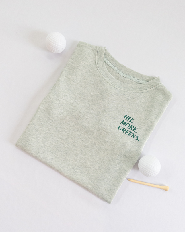 Light green kids golf t-shirt with minimalist text reading 'Hit. More. Greens.' in dark green, displayed flat lay with golf balls and a wooden tee, perfect for young golf enthusiasts.