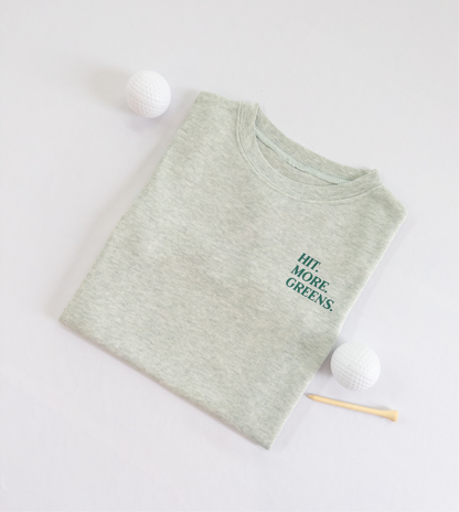 Light green kids golf t-shirt with minimalist text reading 'Hit. More. Greens.' in dark green, displayed flat lay with golf balls and a wooden tee, perfect for young golf enthusiasts.