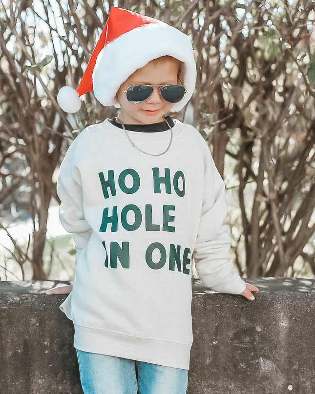Child wearing a Christmas-themed sweater with text, sunglasses, and a Santa hat outdoors.