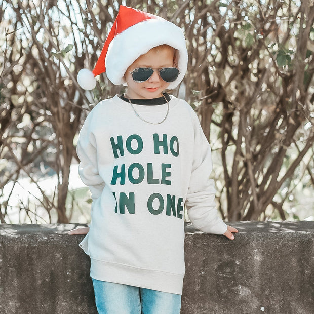 Child wearing a Christmas-themed sweater with text, sunglasses, and a Santa hat outdoors.