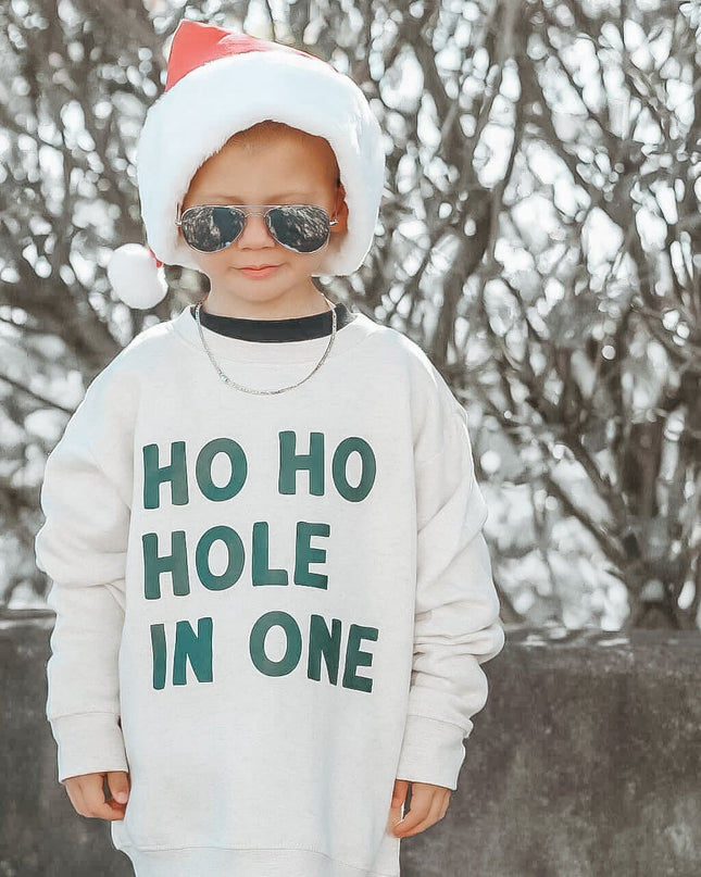 Child wearing a sweater with 'HO HO HOLE IN ONE' text, sunglasses, and a Santa hat outdoors.
