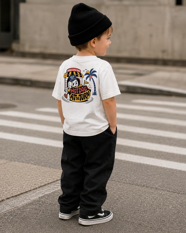 A child standing on a city street crosswalk, viewed from behind, wearing a white t-shirt with a large colorful Adidas Originals graphic on the back, black pants, and a black beanie.