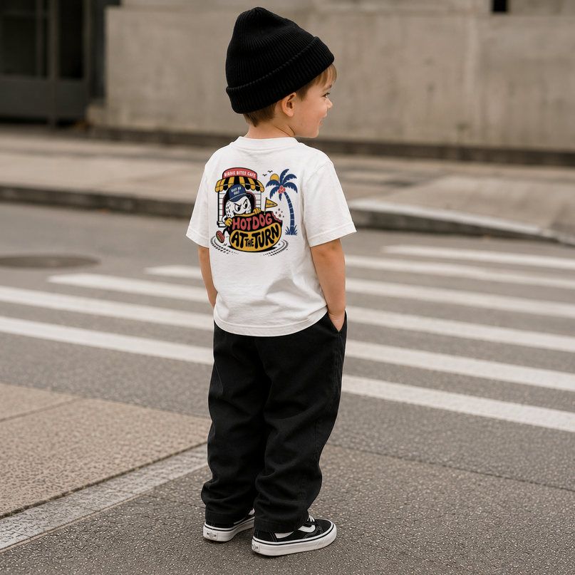 A child standing on a city street crosswalk, viewed from behind, wearing a white t-shirt with a large colorful Adidas Originals graphic on the back, black pants, and a black beanie.