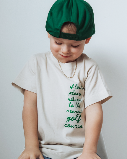 Child wearing a green beret and a t-shirt with text, sitting on a stool against a plain background