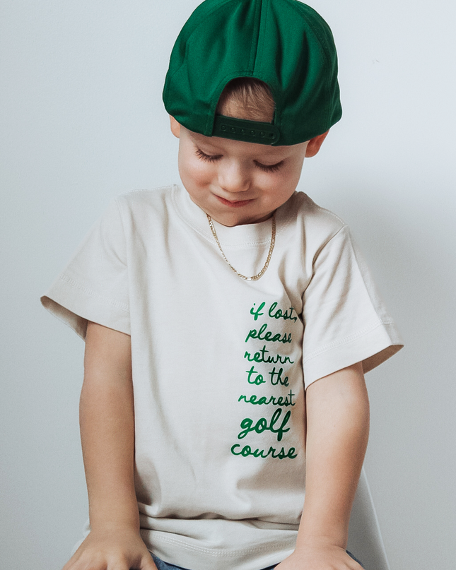 Child wearing a green beret and a t-shirt with text, sitting on a stool against a plain background