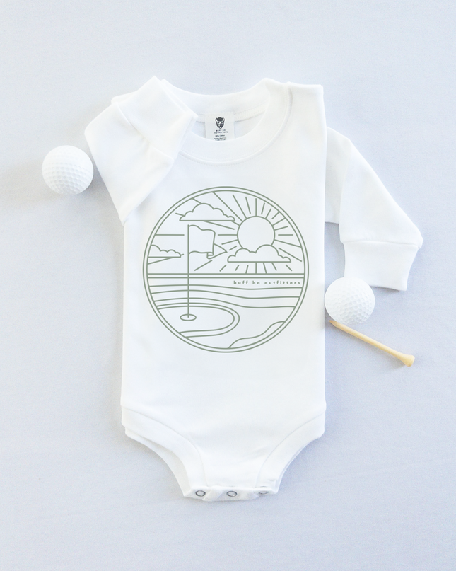 White long-sleeve baby onesie featuring a minimalist golf course design with a flag, sun, and clouds. The onesie is laid flat with two golf balls and a wooden tee beside it.
