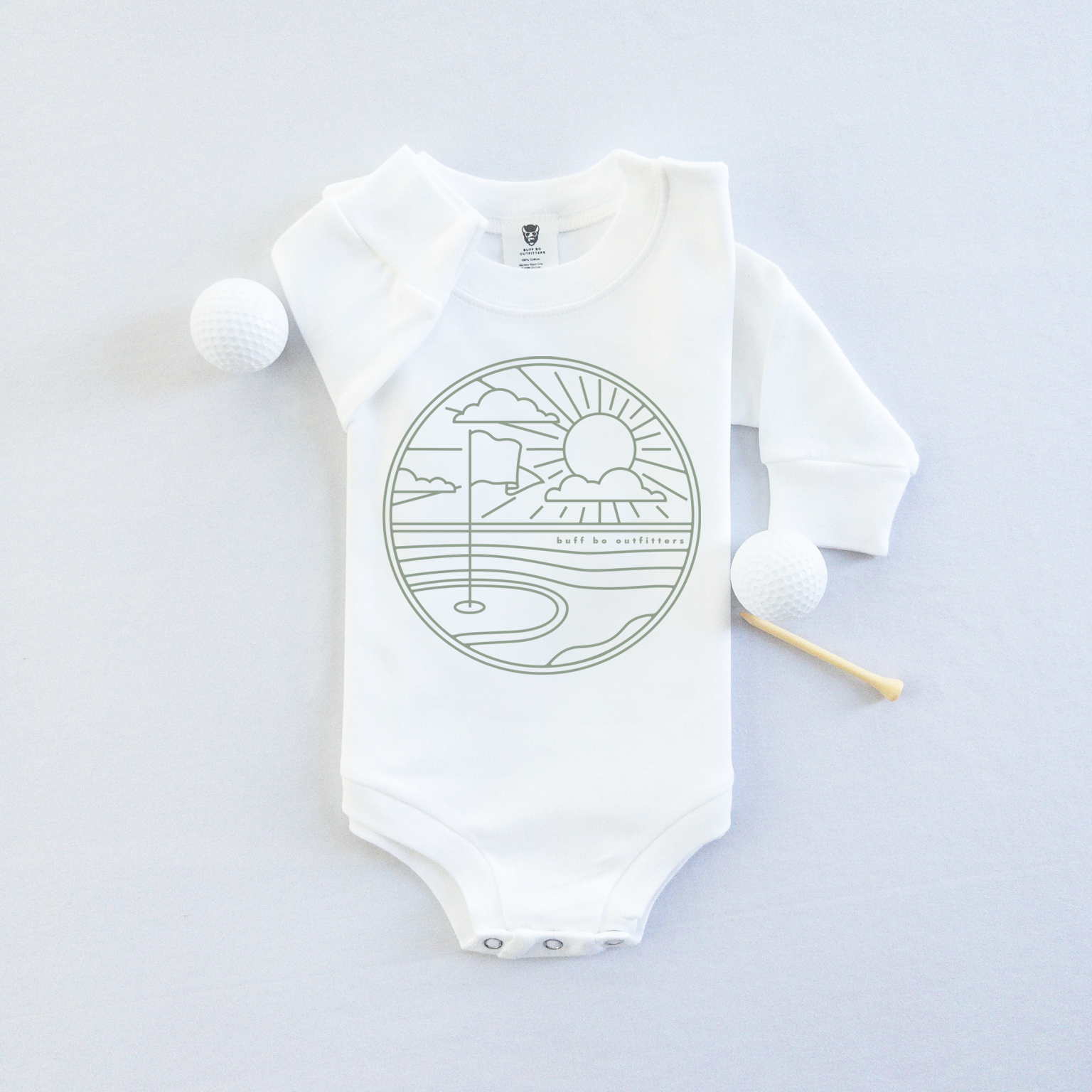 White long-sleeve baby onesie featuring a minimalist golf course design with a flag, sun, and clouds. The onesie is laid flat with two golf balls and a wooden tee beside it.