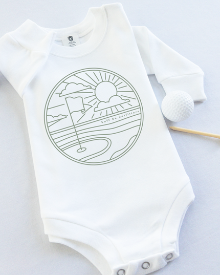 White long-sleeve baby onesie featuring a minimalist golf course design with a flag, sun, and clouds. The onesie is laid flat with two golf balls and a wooden tee beside it.