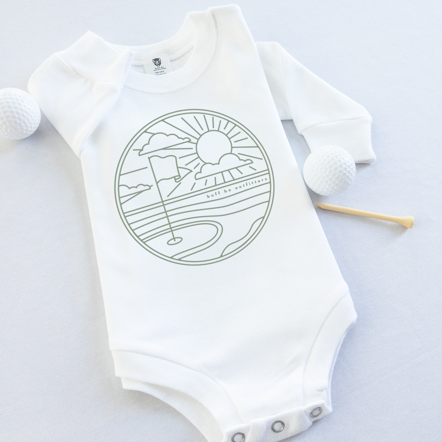 White long-sleeve baby onesie featuring a minimalist golf course design with a flag, sun, and clouds. The onesie is laid flat with two golf balls and a wooden tee beside it.