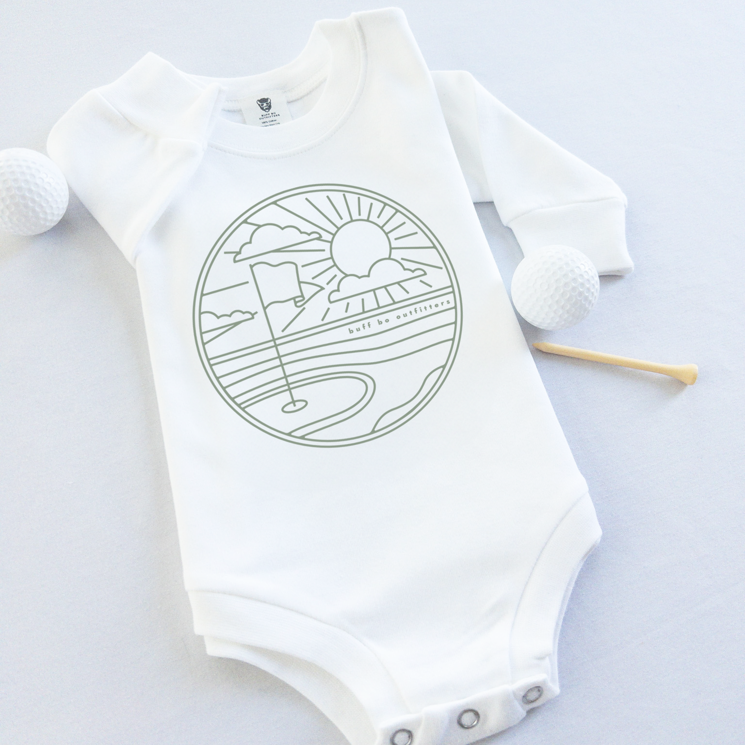 White long-sleeve baby onesie featuring a minimalist golf course design with a flag, sun, and clouds. The onesie is laid flat with two golf balls and a wooden tee beside it.