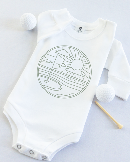 White long-sleeve baby onesie featuring a minimalist golf course design with a flag, sun, and clouds. The onesie is laid flat with two golf balls and a wooden tee beside it.
