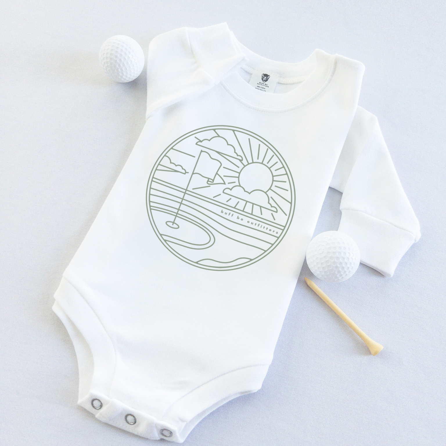 White long-sleeve baby onesie featuring a minimalist golf course design with a flag, sun, and clouds. The onesie is laid flat with two golf balls and a wooden tee beside it.