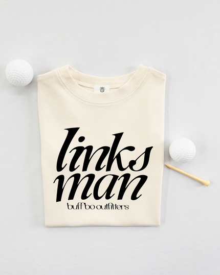 Folded cream-colored kids' t-shirt with bold black text that reads "links man" and "buff bo outfitters" beneath, displayed on a light background with two white golf balls and a wooden tee.
