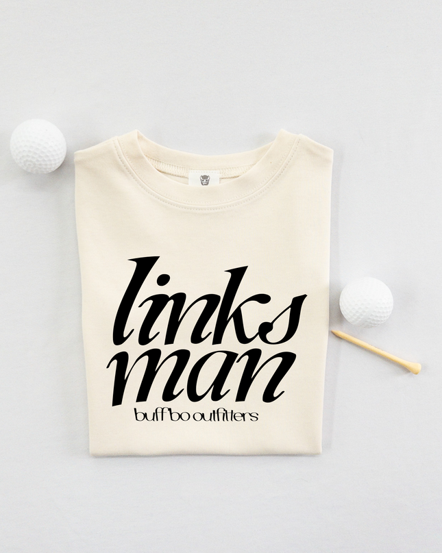 Folded cream-colored kids' t-shirt with bold black text that reads "links man" and "buff bo outfitters" beneath, displayed on a light background with two white golf balls and a wooden tee.
