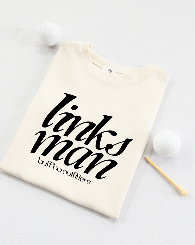 Folded cream-colored kids' t-shirt with bold black text that reads "links man" and "buff bo outfitters" beneath, displayed on a light background with two white golf balls and a wooden tee.
