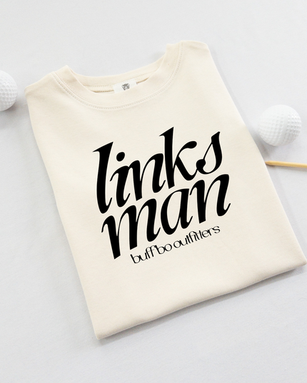 Folded cream-colored kids' t-shirt with bold black text that reads "links man" and "buff bo outfitters" beneath, displayed on a light background with two white golf balls and a wooden tee.
