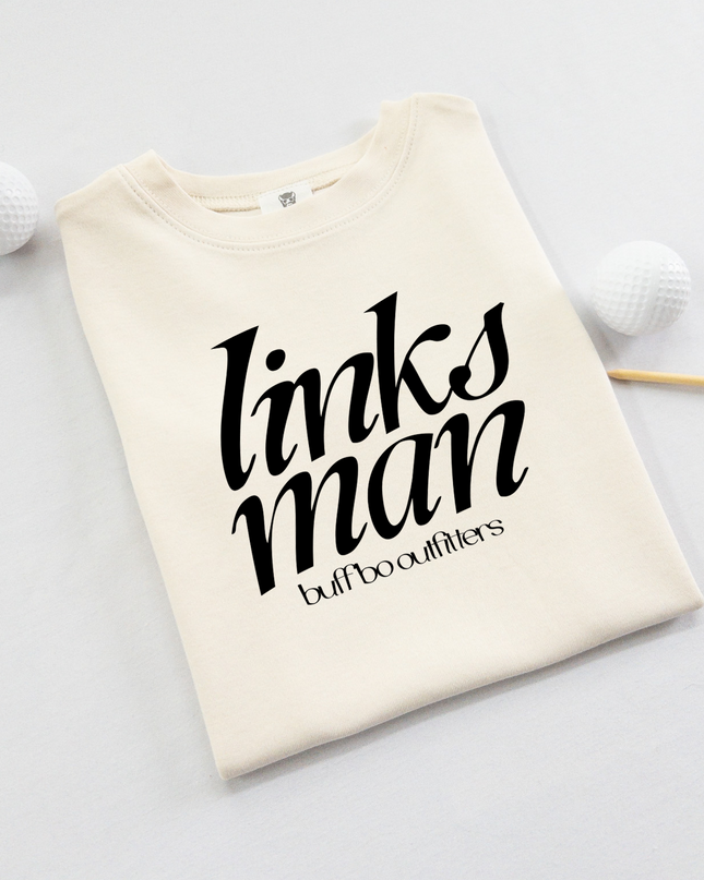 Folded cream-colored kids' t-shirt with bold black text that reads "links man" and "buff bo outfitters" beneath, displayed on a light background with two white golf balls and a wooden tee.
