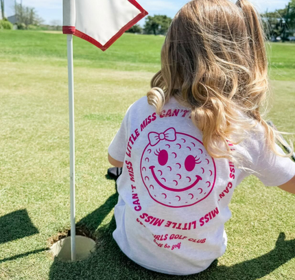 Child wearing a white t-shirt with a smiley face design on a golf course