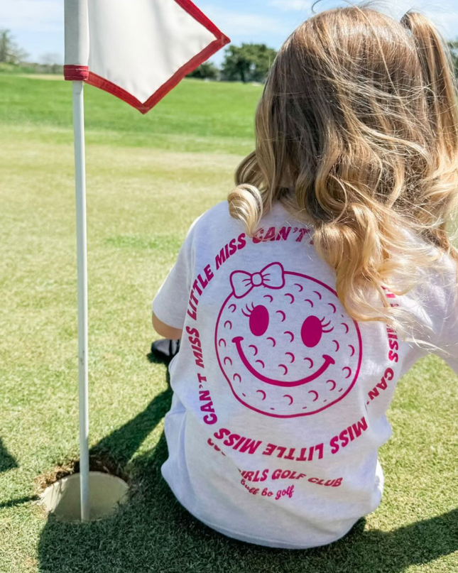 Child wearing a white t-shirt with a smiley face design on a golf course