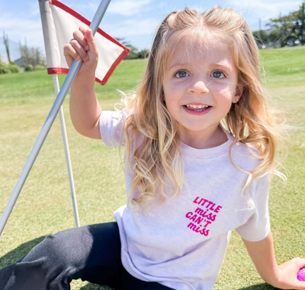 Child sitting on a golf green holding a flag, wearing a t-shirt with text.