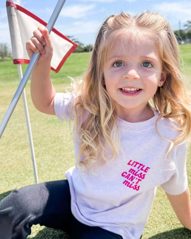 Child sitting on a golf green holding a flag, wearing a t-shirt with text.