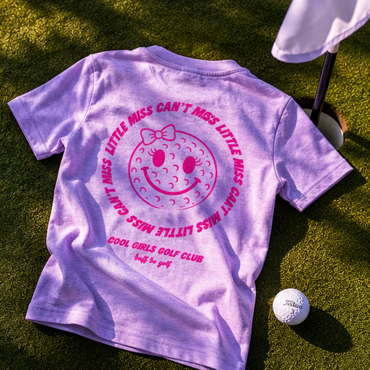Purple t-shirt with pink text and graphics on a grassy surface with a golf ball and club.