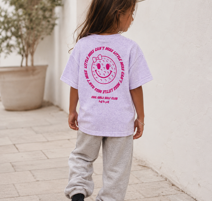 Child wearing a light purple t-shirt with pink text and graphics outdoors.