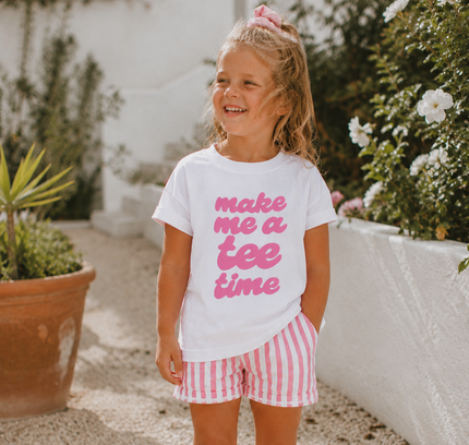 Young girl wearing a white t-shirt with pink text and striped shorts, standing outdoors.