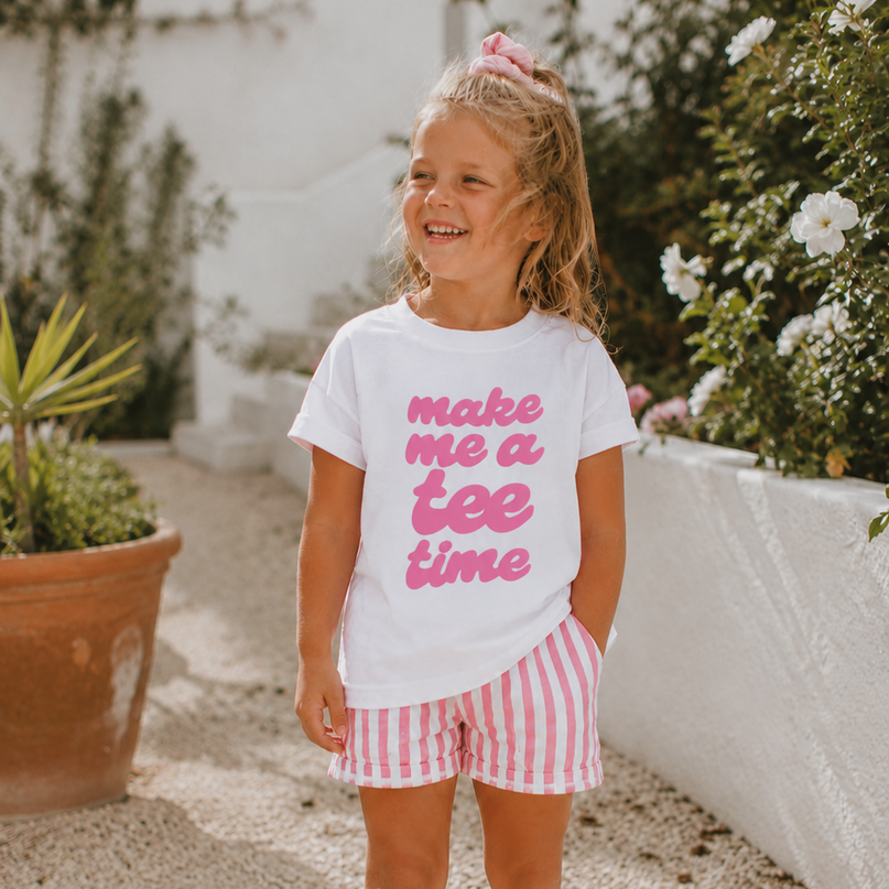 Young girl wearing a white t-shirt with pink text and striped shorts, standing outdoors.