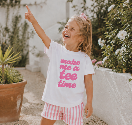 Child wearing a white t-shirt with pink text and red striped shorts, standing outdoors.