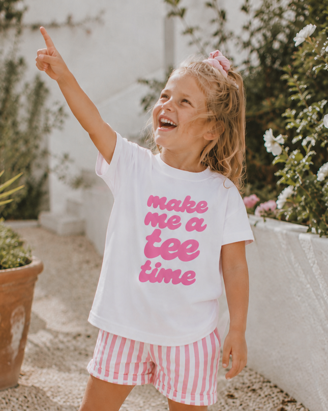 Child wearing a white t-shirt with pink text and red striped shorts, standing outdoors.