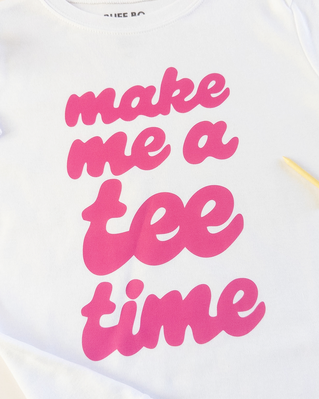 White t-shirt with pink text 'make me a tee time' on a light background