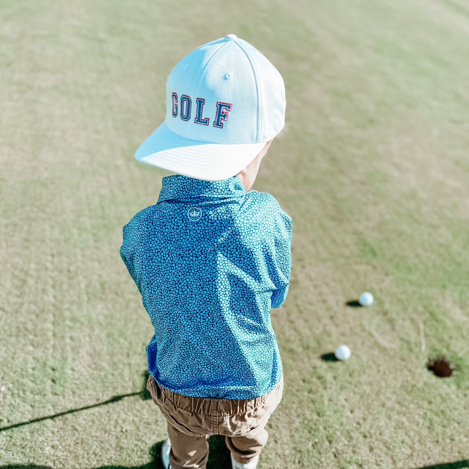 Young child wearing a white "GOLF" embroidered hat and a blue patterned shirt, standing on a golf course near two golf balls and a hole.
